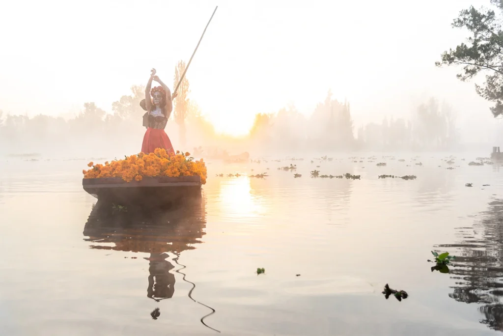 A woman dressed up to resemble La Catrina, riding on a traditional trajinera boat decorated with marigold flowers, during sunrise on the Xochimilco canals in Mexico City.