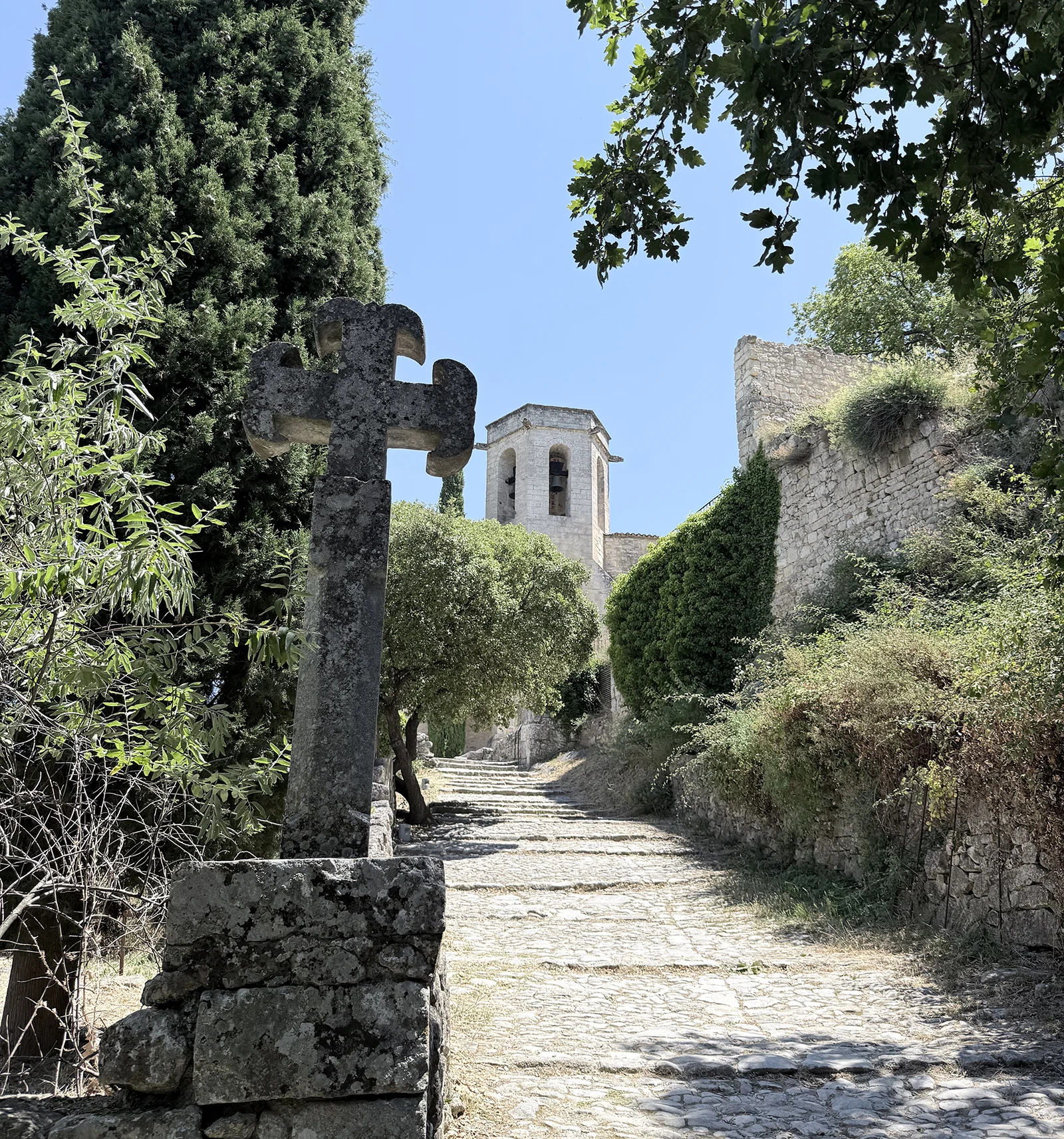 Cobblestone path leading up through Oppède-le-Vieux toward the Romanesque church bell tower, with a weathered stone cross in the foreground and ivy-covered stone walls on either side